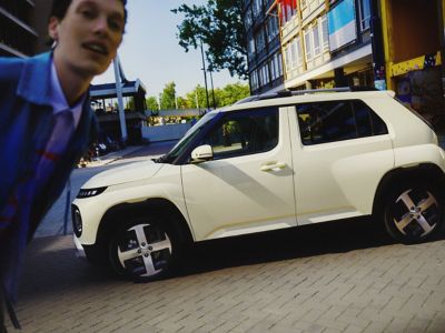 Zijaanzicht van een INSTER Cross die aan de laadpaal hangt terwijl een vrouw tegen de achterzijde leunt