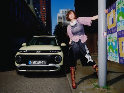 A woman standing next to the new Hyundai INSTER small electric car holding on to a lamp post.