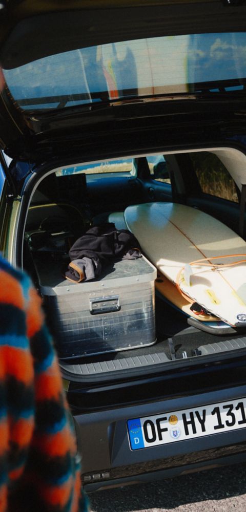 A close-up viw of a man in front of the Hyundai Inster Cross with the trunk open, showcasing its spacious interior with a box and surfboard.