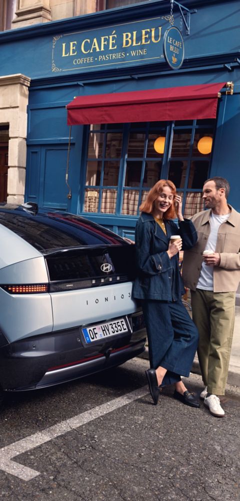 Blue Hyundai IONIQ 3 parked on the street in front of a cafe with two people holding coffee cups.