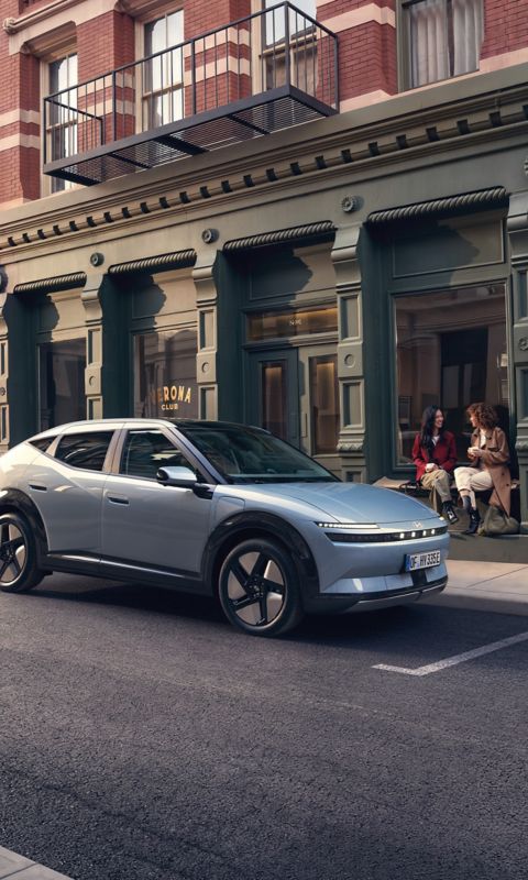 Ein Hyundai IONIQ 3, der auf einer Straße in der Stadt geparkt ist, daneben sitzen zwei Frauen auf einer Treppe.