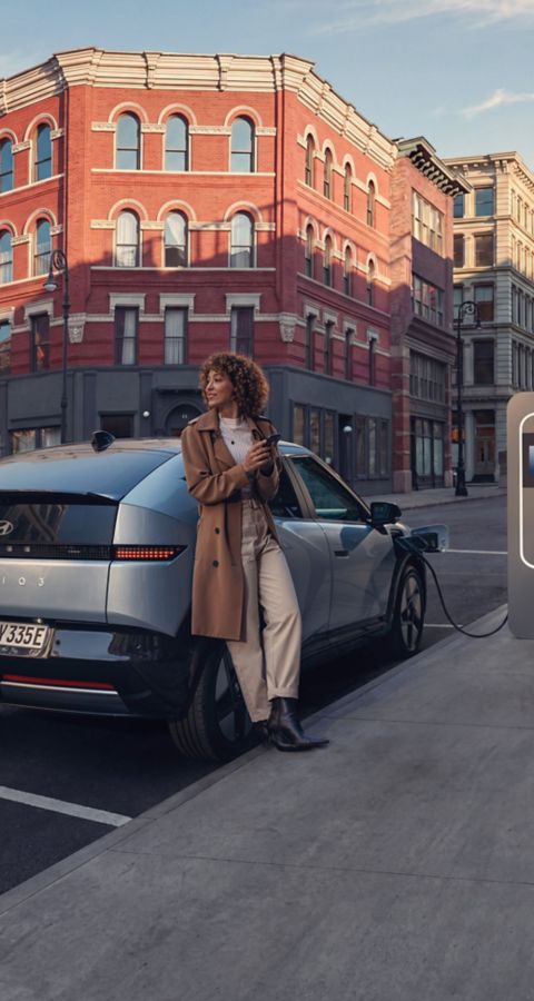 Hyundai IONIQ 3 Prime lädt an einer Ladestation in der Stadt. Eine Frau lehnt an dem Hyundai IONIQ 3 Prime.