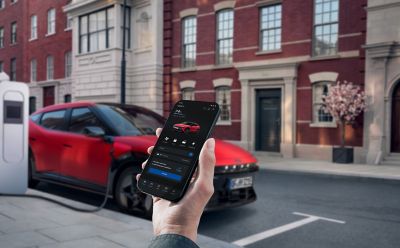 A hand holding a smartphone next to the Hyundai IONIQ 3 while it is plugged in to charge.