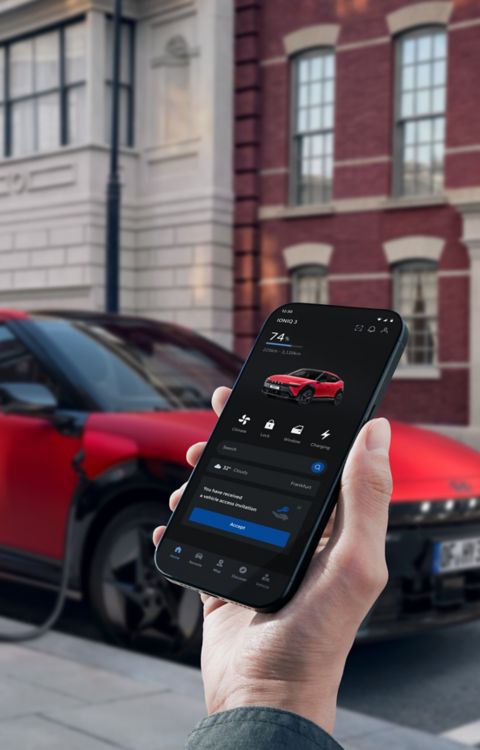 A hand holding a smartphone next to the Hyundai IONIQ 3 while it is plugged in to charge.