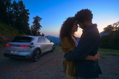 Two people embracing in front of a Hyundai IONIQ 5 being charged, with a scenic twilight backdrop of trees and mountains.