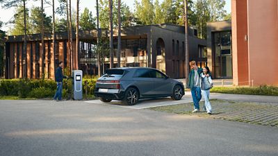 Hyundai IONIQ 5 parked at an outdoor charging station near modern buildings, with one person standing by the charger and two people walking nearby.