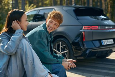 To personer sitter utendørs foran en Hyundai IONIQ 5, omgitt av trær og grøntområder.