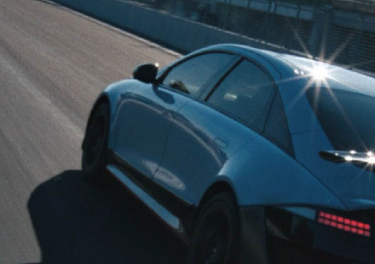 A Hyundai IONIQ 6 N speeds past on the track, reflecting sunlight with a modern building in the background.