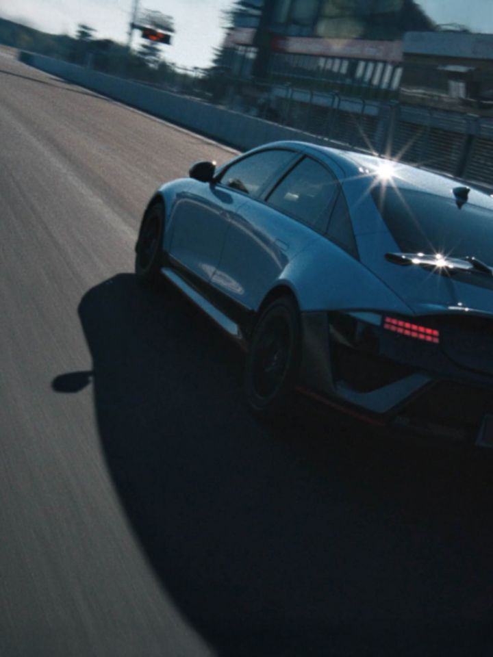 A Hyundai IONIQ 6 N speeds past on the track, reflecting sunlight with a modern building in the background.