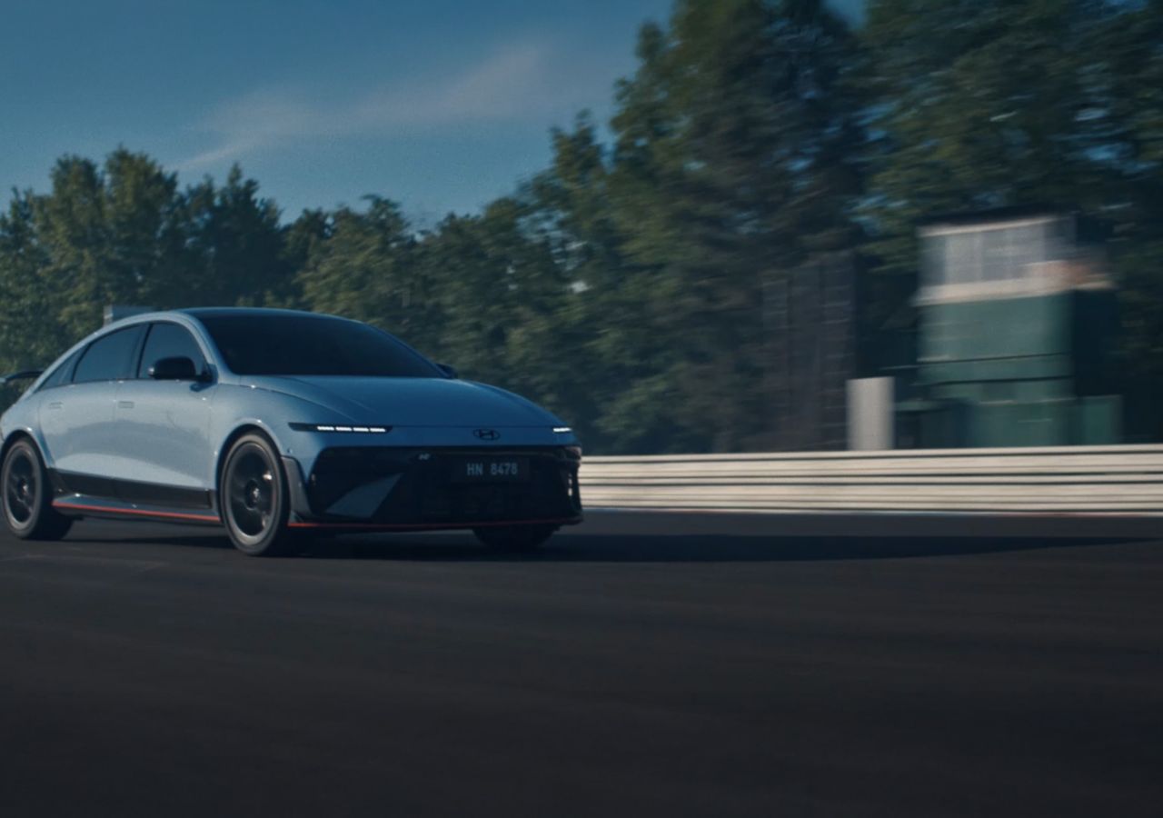 A Hyundai IONIQ 6 N electric performance car speeds along a racetrack with trees and a building in the background.