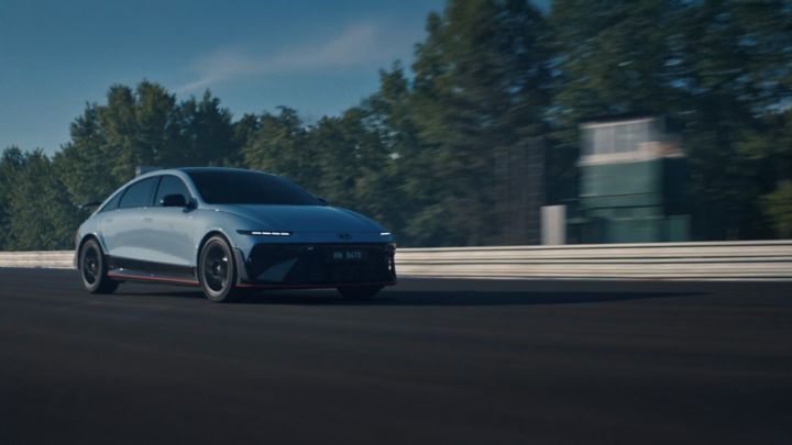 A Hyundai IONIQ 6 N electric performance car speeds along a racetrack with trees and a building in the background.