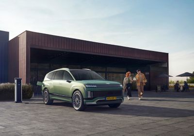 Zicht op geparkeerde Hyundai IONIQ 9 driekwart van voren met 2 mensen rechts ernaast en modern pand op achtergrond.