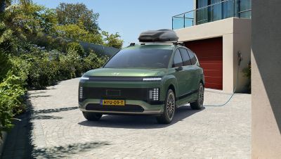 Zicht op Hyundai IONIQ 9 driekwart van voren met dakkoffer op het dak, geparkeerd voor woonhuis en met laadkabel aangesloten op oplaadpunt.