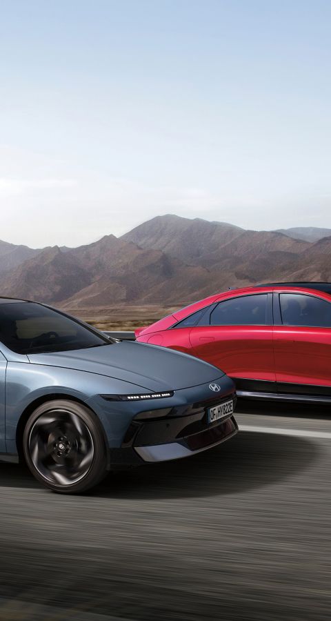 A grey IONIQ 6 and a red IONIQ 6 N Line driving next to each other on a highway with mountains in the background.	