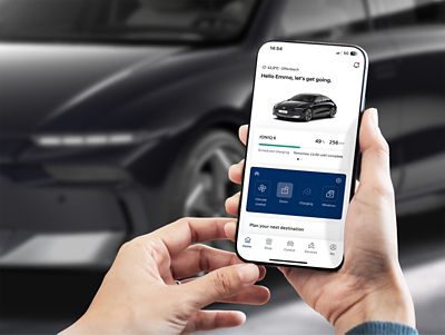 A person is looking at a smartphone with myHyundai app. In background is Hyundai IONIQ 6 parked on the street.