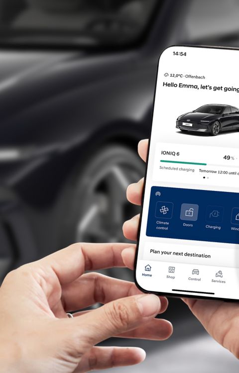 A person is looking at a smartphone with myHyundai app. In background is Hyundai IONIQ 6 parked on the street.