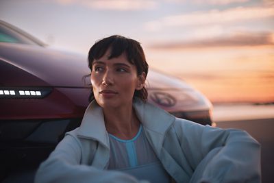A woman sitting and leaning on the front bumper Hyundai IONIQ 6 N Line parked next to the ocean.