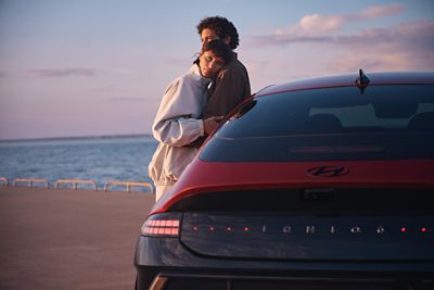 A man and a woman hugging while leaning on a Hyundai IONIQ 6 N Line parked next to the ocean.