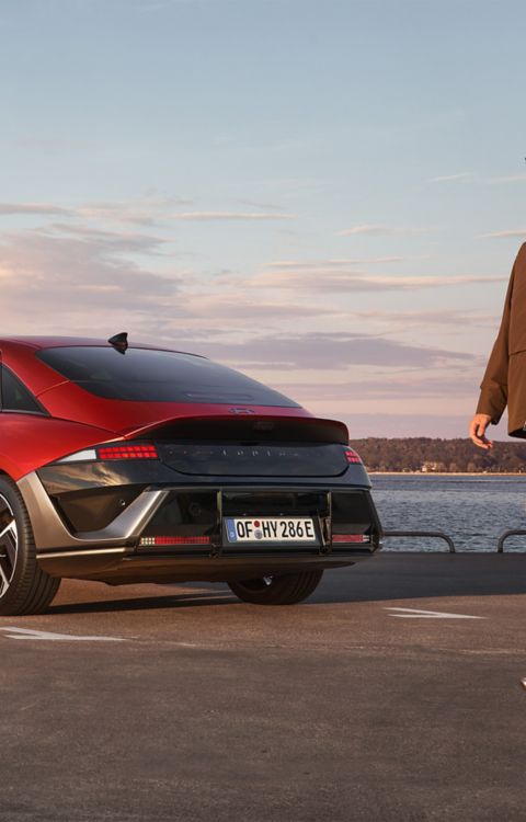 A man and a woman walking past a red Hyundai IONIQ 6 N Line parked next to the ocean.