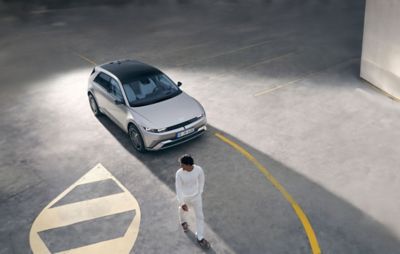 Hyundai IONIQ 6 parked in an empty parking lot with yellow markings, viewed from above. A person wearing white clothing walks nearby.