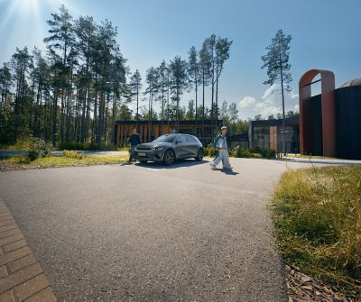 A man stands by a Hyundai IONIQ 5 parked on an asphalt lot, while a couple walks away from the car. In the background are tall trees and modern buildings, all under a sunny sky.