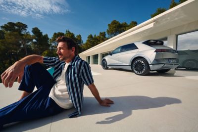 A man sitting in a relaxed pose near the Hyundai IONIQ 5 EV vehicle on a bright, sunny day.