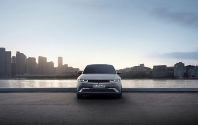 Vista frontal del nuevo Hyundai IONIQ 5 aparcado junto a un río con la ciudad a la luz del atardecer de fondo.