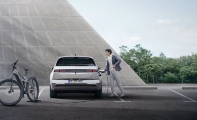 A man using a charging cable to connect an electric bicycle to a Hyundai IONIQ 5 near a pyramid.