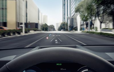 View from the driver’s seat of the Hyundai IONIQ 6, featuring a head-up display showing navigation directions, speed (80 km/h), and traffic information on a clear urban road.