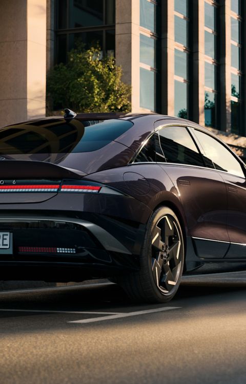 HYUNDAI IONIQ 6 driving through a modern city street, shown from the rear with a sleek aerodynamic design and urban architecture in the background.
