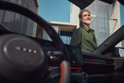 Interior view of a Hyundai KONA EV showing the steering wheel and dashboard, with a bearded man in a green jacket standing outside a modern building.