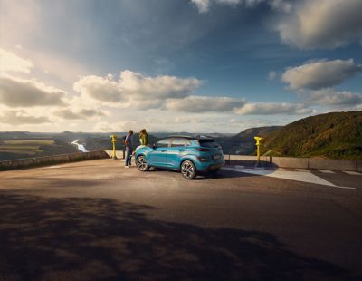 Ein Paar mit ihrem Hyundai KONA Elektro schaut von einem Aussichtspunkt in den Bergen über ein Sonnenbeleuchtetes Tal.