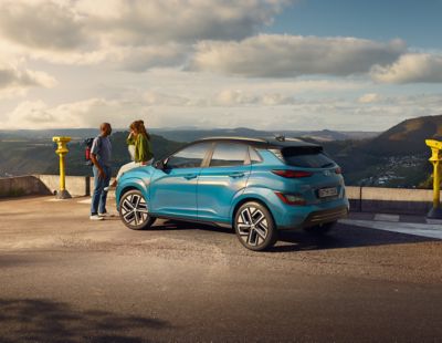 Ein Hyundai KONA Elektro parkt auf einer Aussichtsplattform, eine Frau sitzt auf der Motorhaube, ihr gegenüber steht ein Mann mit Rucksack.