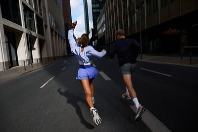 Two runners sprint down an empty urban street between tall office buildings, one arm raised.