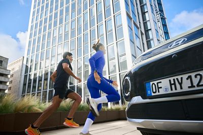 Bir şehir meydanında cam cepheli bir gökdelenin yanında park etmiş bir Hyundai’nin yanından koşarak geçen iki koşucu. 