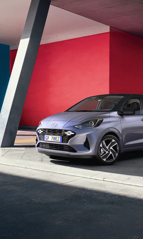 Hyundai i10 hatchback front three-quarter parked under a modern overhang with bold red and blue walls.