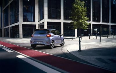 Hyundai i10 seen from rear three-quarter turning on a quiet city street in front of modern office buildings.
