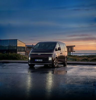 Driekwart voorzijde Hyundai STARIA Electric Passenger met op achtergrond een zonsondergang..
