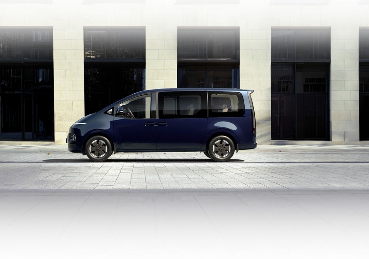 Seitenansicht des Hyundai STARIA Electric, der vor einem modernen Gebäude geparkt ist, wobei sein elegantes Design, die grossen Fenster und die Schiebetüren hervorgehoben werden.