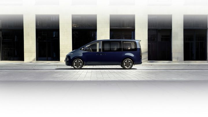 Side view of Hyundai STARIA Electric parked in front of a modern building, highlighting its sleek design, large windows, and sliding doors.