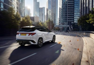 Witte Hyundai TUCSON N Line rijdt door een moderne stad met hoge kantoorgebouwen, terwijl herfstbladeren over de weg waaien in het ochtendlicht.