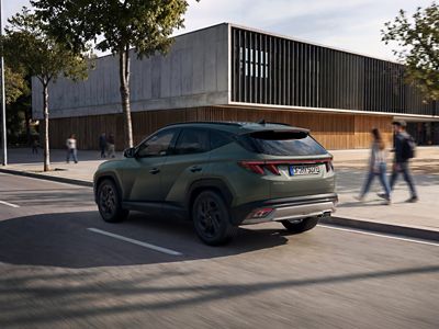 Pohled zezadu na Hyundai TUCSON jedoucí po ulici s moderní budovou, stromy a chodci v pozadí.
