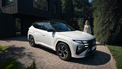 Hyundai TUCSON N Line zaparkovaný před domem, vedle kterého stojí žena.