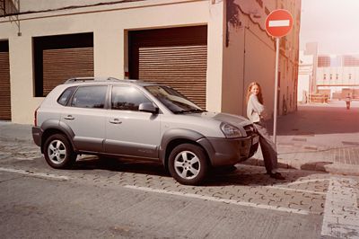 A retro photo of a 2005 Hyundai TUCSON parked by a building with a woman leaning on the bonnet.	