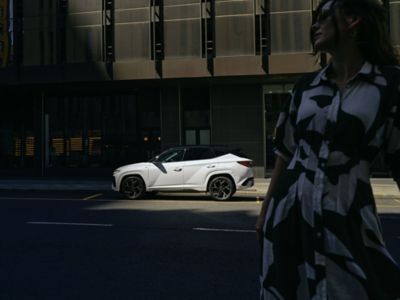 Der Hyundai TUCSON parkt ein einer Straße, im Vordergrund steht eine Frau mit Sonnenbrille.