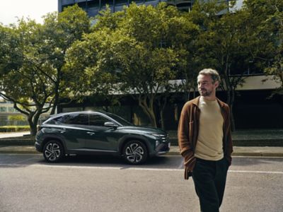 A man walking near a Hyundai TUCSON Hybrid parked by some trees.