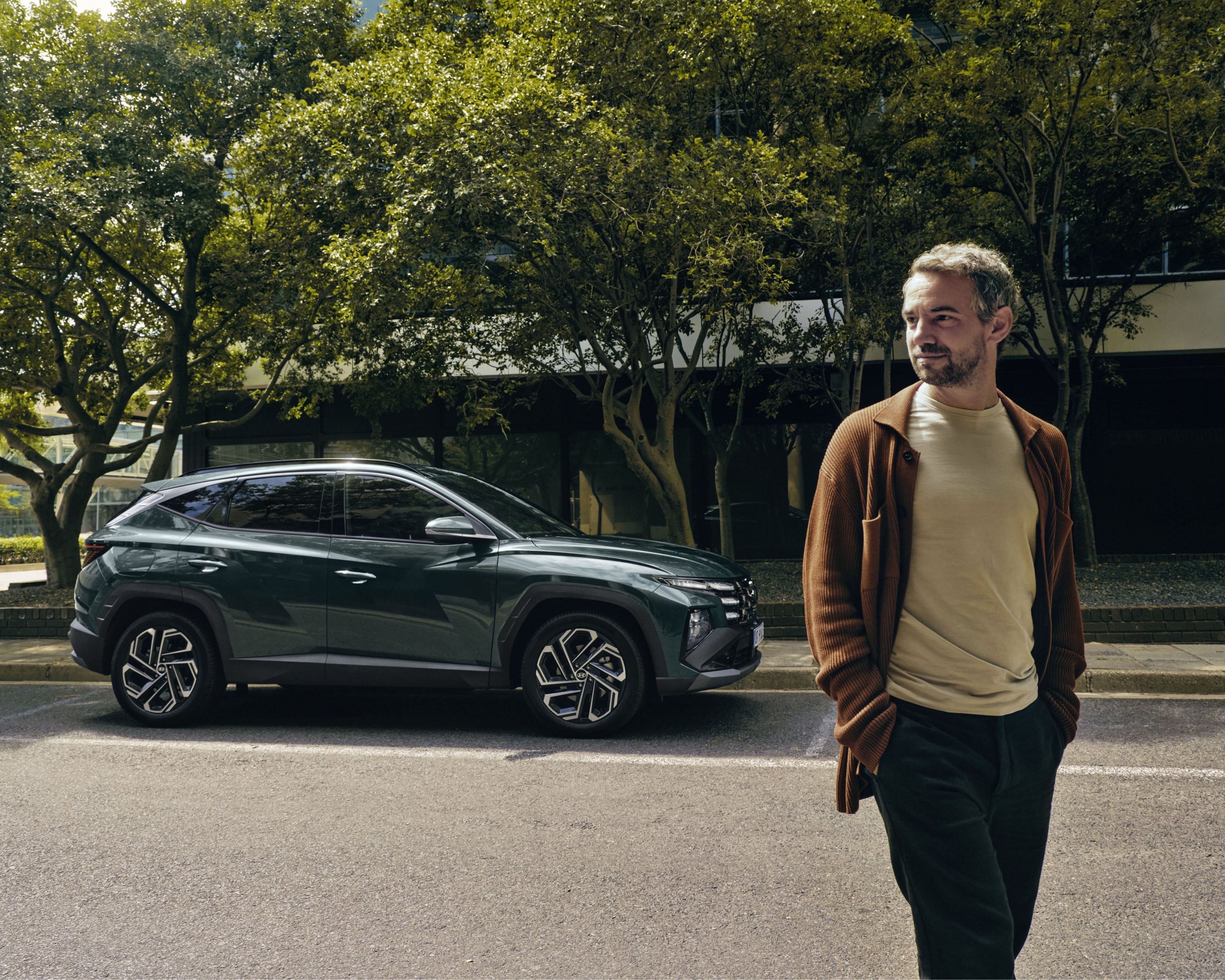 A man walking near a Hyundai TUCSON Hybrid parked by some trees.