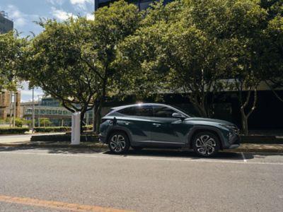 The Hyundai TUCSON Plug-in Hybrid plugged in to charge on a street under some trees. 