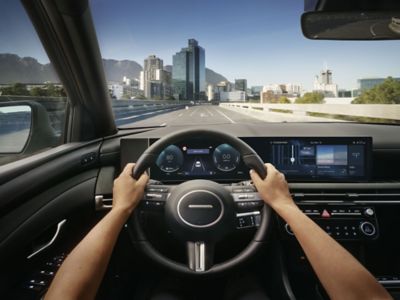 Twee armen aan stuur in cockpit Hyundai TUCSON met uitzicht op weg en gebouwen op achtergrond.