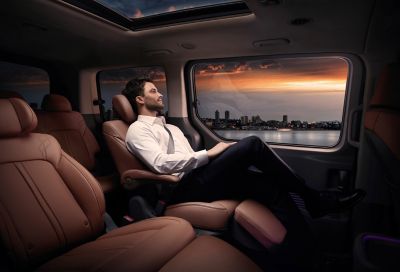 Ein Mann genießt im Relax-Sitz eines Hyundai STARIA den Ausblick auf eine Skyline bei Sonnenuntergang. 